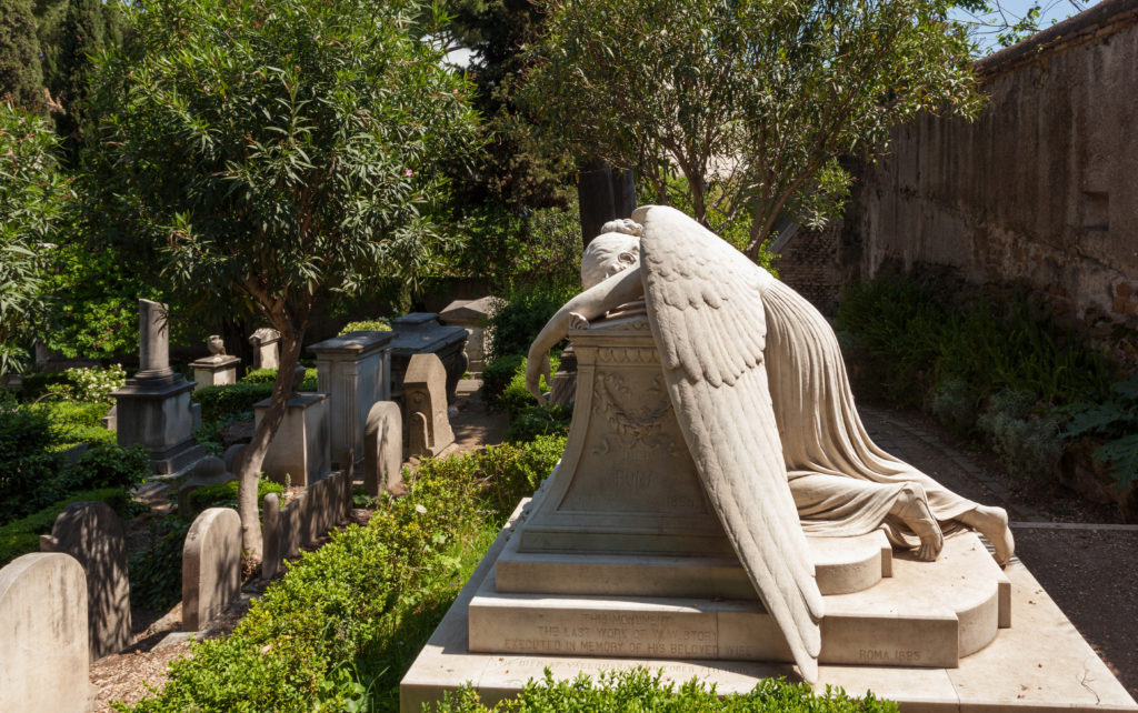 The Weeping Angle statue in Rome's Cimitero Acattolico (aka "The Protestant Cemetery"). It shows a marble-carved angel folded over a grave weeping. Her wings drape beautifully to the ground. Other graves and part of the ancient city wall remain visible behind her. The 1894 sculpture was created by William Wetmore Story for his wife Evelyn Story.