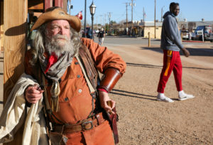 A street scene in Tombstone, Arizona. A re-enactor dressed as a prospector leans against a hitching point, while a tourist in athletic gear does a double-take