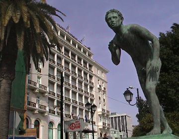 A verigris-bronze statue appears ready to run in front of the Grande Bretagne hotel before its early-millennia fact-lift. The white building looks a bit soot-stained and shabby. A palm free stands to the side.